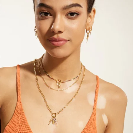 A model wearing a variety of Ania Haie jewelry from the Pop Charms Collection including earrings and necklaces with a spaghetti strap orange top