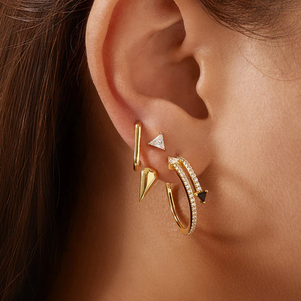 The 14K Yellow Gold Plated Large Oval Huggie Hoop Earrings being worn by a model and stacked with other hoop and stud earrings